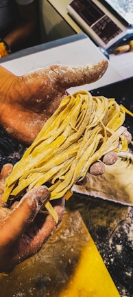 A person with flour-dusted hands is holding a bunch of freshly made pasta. The background shows a countertop sprinkled with flour and various kitchen tools.