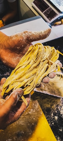 A person with flour-dusted hands is holding a bunch of freshly made pasta. The background shows a countertop sprinkled with flour and various kitchen tools.