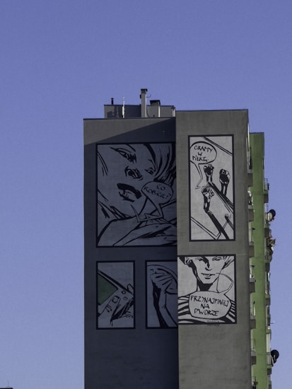 A large building facade displaying multiple comic-style images. The images feature detailed, monochromatic illustrations including a woman's face with a speech bubble, two people playing ball with the text 'GRAMY W PILKE,' and a smiling person with text below saying 'PRZYNAJMNIEJ NA DWORZE.' The wall and images are primarily in black and white tones, set against a clear blue sky.