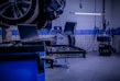 A car service or repair garage is depicted, with a vehicle hoisted on a lift. A laptop is placed on a nearby workstation with an open drawer filled with tools. The environment features tiled walls and floor, and equipment such as a tire changer and a balancing machine. Fluorescent lighting provides illumination.