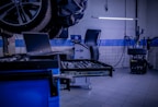 A car service or repair garage is depicted, with a vehicle hoisted on a lift. A laptop is placed on a nearby workstation with an open drawer filled with tools. The environment features tiled walls and floor, and equipment such as a tire changer and a balancing machine. Fluorescent lighting provides illumination.