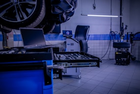 A car service or repair garage is depicted, with a vehicle hoisted on a lift. A laptop is placed on a nearby workstation with an open drawer filled with tools. The environment features tiled walls and floor, and equipment such as a tire changer and a balancing machine. Fluorescent lighting provides illumination.