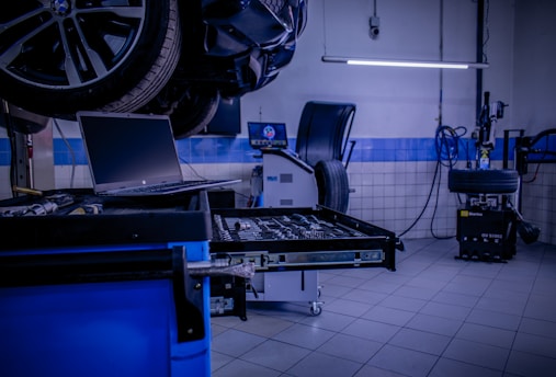 A car service or repair garage is depicted, with a vehicle hoisted on a lift. A laptop is placed on a nearby workstation with an open drawer filled with tools. The environment features tiled walls and floor, and equipment such as a tire changer and a balancing machine. Fluorescent lighting provides illumination.