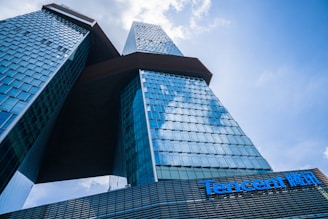A modern skyscraper with a unique architectural design featuring two interconnected towers. The building's facade is made of blue-tinted glass reflecting the sky, and it appears massive and imposing from the low-angle perspective. The Tencent logo is prominently displayed on the lower section of the structure.