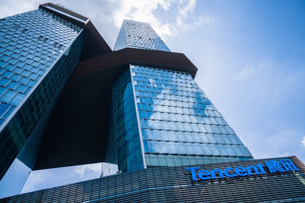 A modern skyscraper with a unique architectural design featuring two interconnected towers. The building's facade is made of blue-tinted glass reflecting the sky, and it appears massive and imposing from the low-angle perspective. The Tencent logo is prominently displayed on the lower section of the structure.