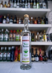 A glass bottle of mezcal with a cork top prominently displayed in the foreground. The label includes the text 'De Maguey Mezcalero, Doble Destilado', and features a green maguey plant illustration. The background shows a blurred array of various liquor bottles lined up on shelves.