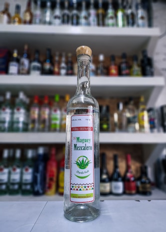 Close-up of a transparent glass bottle labeled 'xa mezcal artesanal' against the agave field.