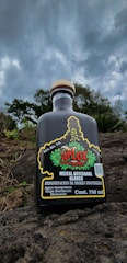 A bottle of mezcal is positioned on rocky terrain with cloudy skies above. The label features vibrant red and green colors with detailed designs, indicating its artisanal craftsmanship.