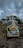 A bottle of mezcal is positioned on rocky terrain with cloudy skies above. The label features vibrant red and green colors with detailed designs, indicating its artisanal craftsmanship.