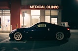 A sleek black sports car is parked outside a medical clinic at night. The building is illuminated by a red neon sign that reads 'MEDICAL CLINIC'. The environment appears quiet and empty, with a dimly lit parking area and subtle reflections on the car's surface.