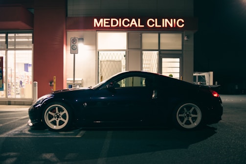 A sleek black sports car is parked outside a medical clinic at night. The building is illuminated by a red neon sign that reads 'MEDICAL CLINIC'. The environment appears quiet and empty, with a dimly lit parking area and subtle reflections on the car's surface.