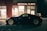 A sleek black sports car is parked outside a medical clinic at night. The building is illuminated by a red neon sign that reads 'MEDICAL CLINIC'. The environment appears quiet and empty, with a dimly lit parking area and subtle reflections on the car's surface.