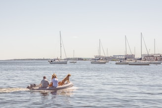 A small inflatable boat is carrying two people and a dog across a calm body of water. In the background, several sailboats are anchored near a distant shore with scattered buildings.