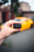 A hand is holding a small black device labeled 'BlueDriver,' with a blurred background that includes a bright yellow sports car.