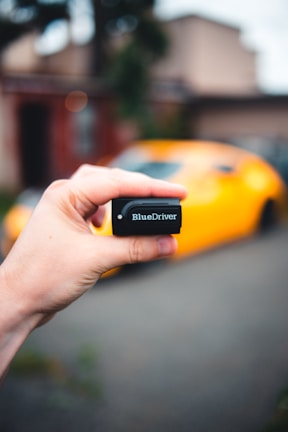 A hand is holding a small black device labeled 'BlueDriver,' with a blurred background that includes a bright yellow sports car.