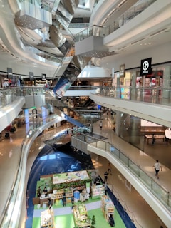 A spacious multi-level shopping mall with sleek, modern architecture. The interior features several floors connected by shiny, metallic escalators. Various high-end retail stores are visible, showcasing popular brand logos. The ground floor hosts a colorful market stall area with people browsing different stalls. Natural light streams in through overhead windows, enhancing the bright and airy atmosphere.