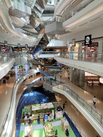 A spacious multi-level shopping mall with sleek, modern architecture. The interior features several floors connected by shiny, metallic escalators. Various high-end retail stores are visible, showcasing popular brand logos. The ground floor hosts a colorful market stall area with people browsing different stalls. Natural light streams in through overhead windows, enhancing the bright and airy atmosphere.