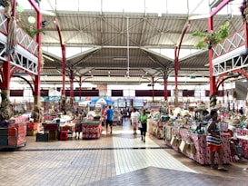 A vibrant indoor market with various stalls displaying merchandise and produce. The market has a high ceiling with red metal supports and plenty of natural light streaming through the windows. Several people are walking and browsing the stalls, which are covered with colorful fabrics.