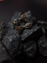 A pair of intricately designed metallic earrings are placed on a pile of dark, textured rocks. The earrings have a vintage look with detailed engravings, adding an element of elegance.