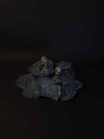 A pair of ornate earrings rests on a pile of dark rocks. The background is black, enhancing the earrings' intricate design and metallic sheen. The earrings have a teardrop shape with detailed embellishments.
