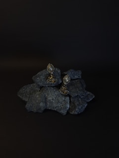 A pair of ornate earrings rests on a pile of dark rocks. The background is black, enhancing the earrings' intricate design and metallic sheen. The earrings have a teardrop shape with detailed embellishments.