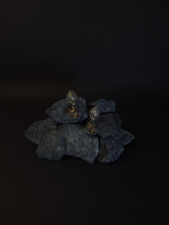 A pair of ornate earrings rests on a pile of dark rocks. The background is black, enhancing the earrings' intricate design and metallic sheen. The earrings have a teardrop shape with detailed embellishments.