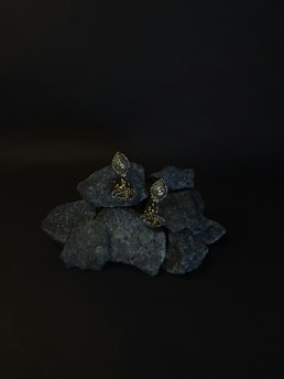 A pair of ornate earrings rests on a pile of dark rocks. The background is black, enhancing the earrings' intricate design and metallic sheen. The earrings have a teardrop shape with detailed embellishments.