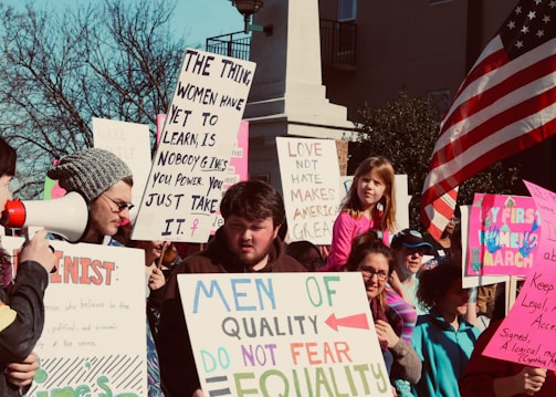 A crowd of people participating in a protest or rally, holding various signs advocating for gender equality and social justice. An American flag is visible in the background.