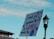 A protest sign is held up against a clear blue sky, featuring the words 'I march for oppressed WOMEN around the world' with decorative elements including a female gender symbol, flowers, and a globe. The background shows part of a building and a street lamp.