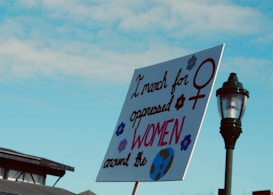 A protest sign is held up against a clear blue sky, featuring the words 'I march for oppressed WOMEN around the world' with decorative elements including a female gender symbol, flowers, and a globe. The background shows part of a building and a street lamp.