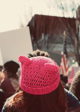 A person wearing a bright pink, knitted hat stands in a crowd. The hat is designed with two pointed cat-ear shapes. Several people are visible in the background, holding signs. There is an American flag to the right side of the image. Trees and a building are visible in the distance.
