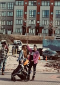 A woman with a raised fist stands beside another woman pushing a stroller with a child in it. They are outdoors in an urban environment, with a backdrop of colorful buildings and parked cars. The people are dressed casually, with the woman raising her fist wearing a pink checkered shirt.