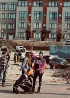 A woman with a raised fist stands beside another woman pushing a stroller with a child in it. They are outdoors in an urban environment, with a backdrop of colorful buildings and parked cars. The people are dressed casually, with the woman raising her fist wearing a pink checkered shirt.