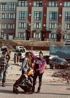 A woman with a raised fist stands beside another woman pushing a stroller with a child in it. They are outdoors in an urban environment, with a backdrop of colorful buildings and parked cars. The people are dressed casually, with the woman raising her fist wearing a pink checkered shirt.