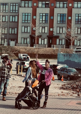 A woman with a raised fist stands beside another woman pushing a stroller with a child in it. They are outdoors in an urban environment, with a backdrop of colorful buildings and parked cars. The people are dressed casually, with the woman raising her fist wearing a pink checkered shirt.
