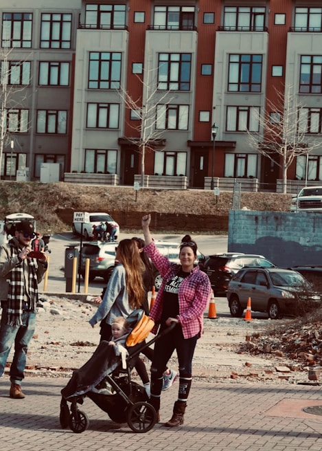 A woman with a raised fist stands beside another woman pushing a stroller with a child in it. They are outdoors in an urban environment, with a backdrop of colorful buildings and parked cars. The people are dressed casually, with the woman raising her fist wearing a pink checkered shirt.