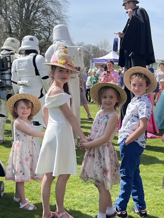 A group of children, dressed in summery outfits and straw hats, stand on a grassy field. They are holding hands and smiling, surrounded by people in costumes including stormtroopers and other characters. There is a man on stilts dressed in black and numerous people in the background, some taking photos.