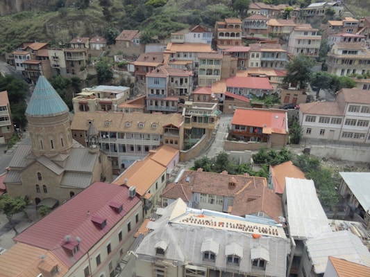 The image captures a picturesque view of a hillside town with traditional buildings featuring red-tiled roofs. A prominent feature is the church with a tall, pointed dome, surrounded by a cluster of tightly packed houses. The scene is set against a backdrop of lush greenery and rugged hills.