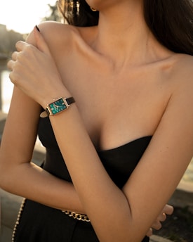 A person is wearing a strapless black outfit, accessorized with a chain belt. They are displaying a stylish wristwatch with a teal face and black leather strap, positioned prominently on the wrist. The surroundings appear sunlit, contributing a warm glow to the skin tones.