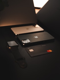 A dark-themed composition feature a smartphone, smartwatch, and a credit card. Each item displays a sleek, modern design, with the phone and smartwatch exhibiting metallic accents, while the credit card has a black matte finish with a prominent MasterCard logo.