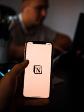 A person holds a smartphone with the Notion logo on the screen. The background is dimly lit, with a blurred figure sitting at a desk possibly using a laptop.