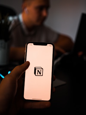 A person holds a smartphone with the Notion logo on the screen. The background is dimly lit, with a blurred figure sitting at a desk possibly using a laptop.
