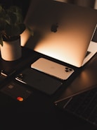 A sleek workspace setup with a laptop displaying an illuminated logo, two smartphones, a smartwatch, and a bank card arranged neatly. A potted plant adds a touch of nature, and the warm lighting creates a cozy atmosphere.