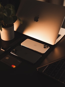 A sleek workspace setup with a laptop displaying an illuminated logo, two smartphones, a smartwatch, and a bank card arranged neatly. A potted plant adds a touch of nature, and the warm lighting creates a cozy atmosphere.