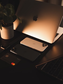 A sleek workspace setup with a laptop displaying an illuminated logo, two smartphones, a smartwatch, and a bank card arranged neatly. A potted plant adds a touch of nature, and the warm lighting creates a cozy atmosphere.