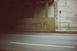 A concrete barrier under an overpass with graffiti saying 'no cops' in black paint. The background includes a signpost, some greenery, and brick and concrete walls.