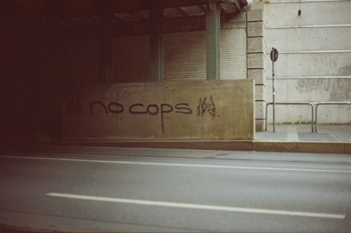 A concrete barrier under an overpass with graffiti saying 'no cops' in black paint. The background includes a signpost, some greenery, and brick and concrete walls.