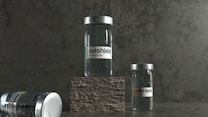 Three vials labeled 'Covishield Covid-19 Vaccine' are positioned against a dark, textured background. Two vials stand upright, with one on a raised stone-like platform, while another lies on its side.