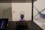 A small electronic device with a digital display is housed under a clear glass dome, mounted on a black and gray stand. The display shows a pixelated icon. The setup appears to be on a shelf alongside a blurred electronic screen on the left. The background includes an empty wall with a subtle chair outline on the right.