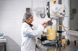 A person dressed in a lab coat and hairnet is working in a laboratory environment. They are using a large beaker filled with a yellow liquid, placed on an electronic balance. The individual is adding an ingredient using a small cup while monitoring the process.